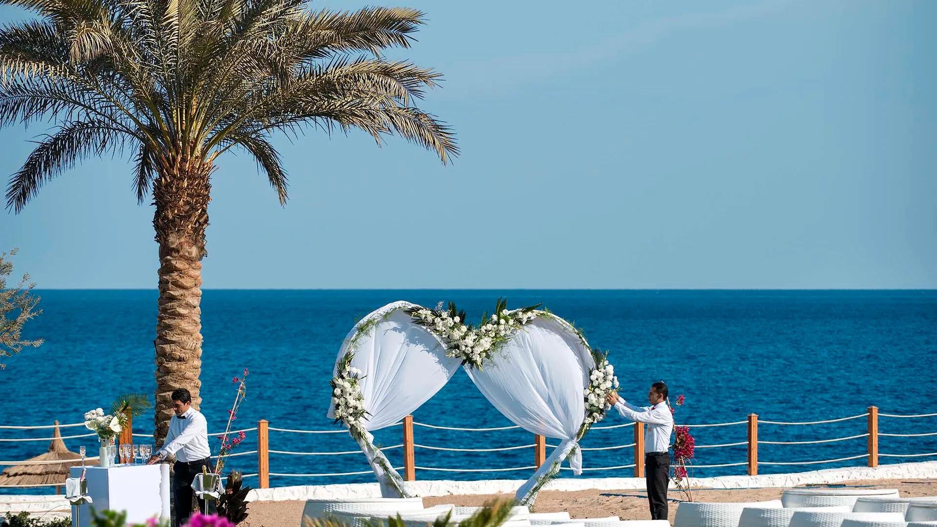 A Couple Of People Standing Next To A Palm Tree And A Large White Umbrella