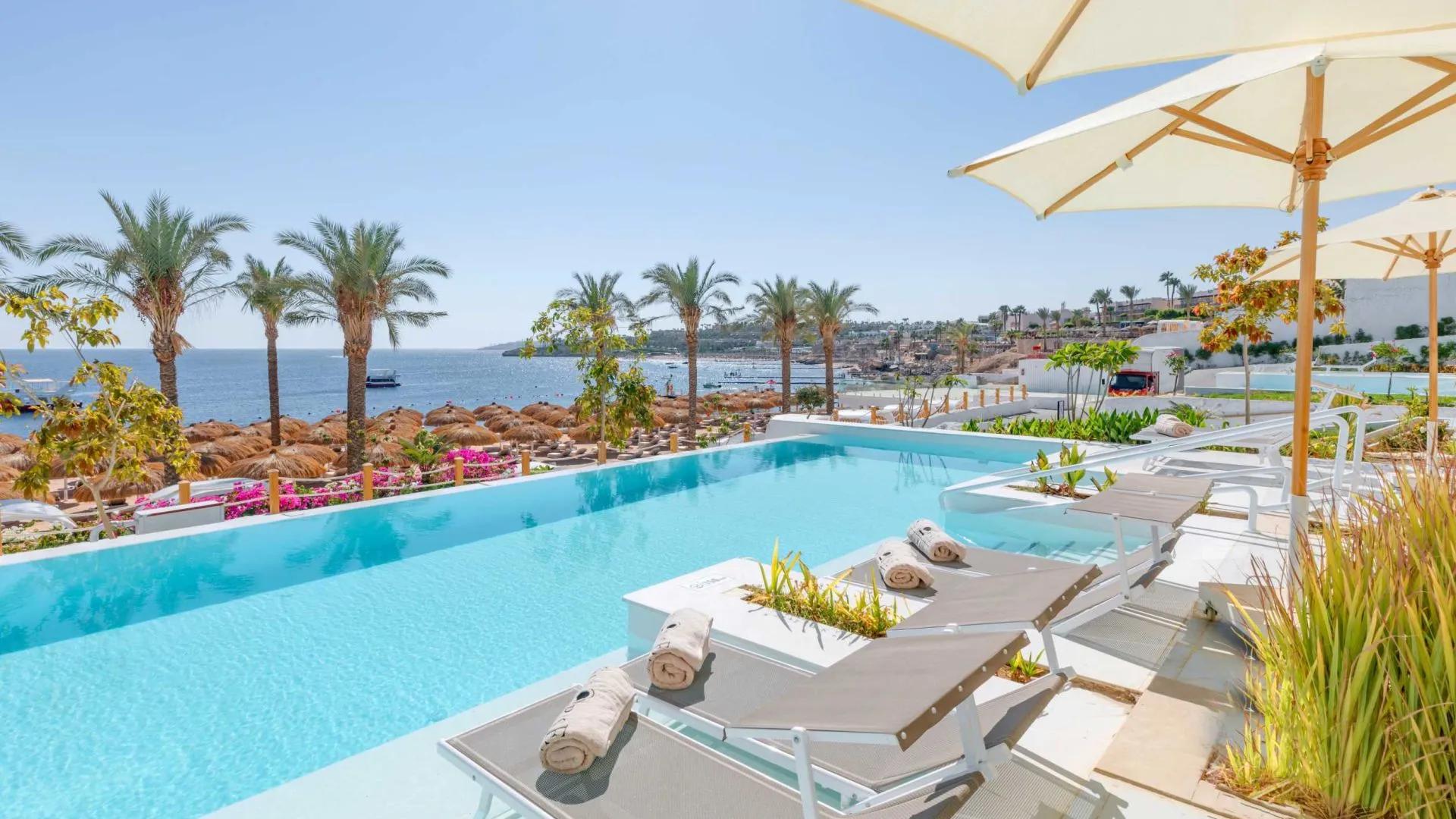 A Pool With Lounge Chairs And Umbrellas By A Beach